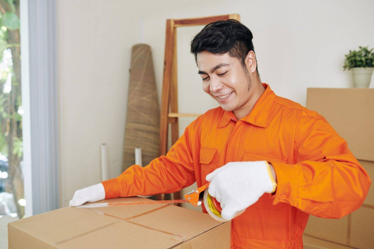 Agent de déménagement en combinaison orange, scotchant des boîtes, ambiance atelier intérieur et cartonage. commentaire: comment bien organiser son déménagement en 1 mois.