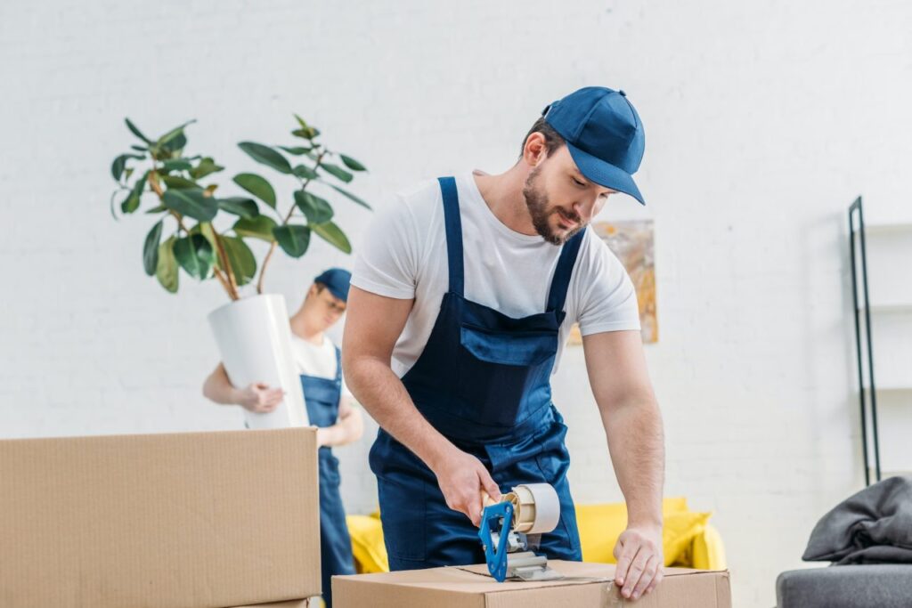 Homme en tenue de travail scelle une boîte avec une sceller et une scie dans un déménagement, intérieur lumineux.