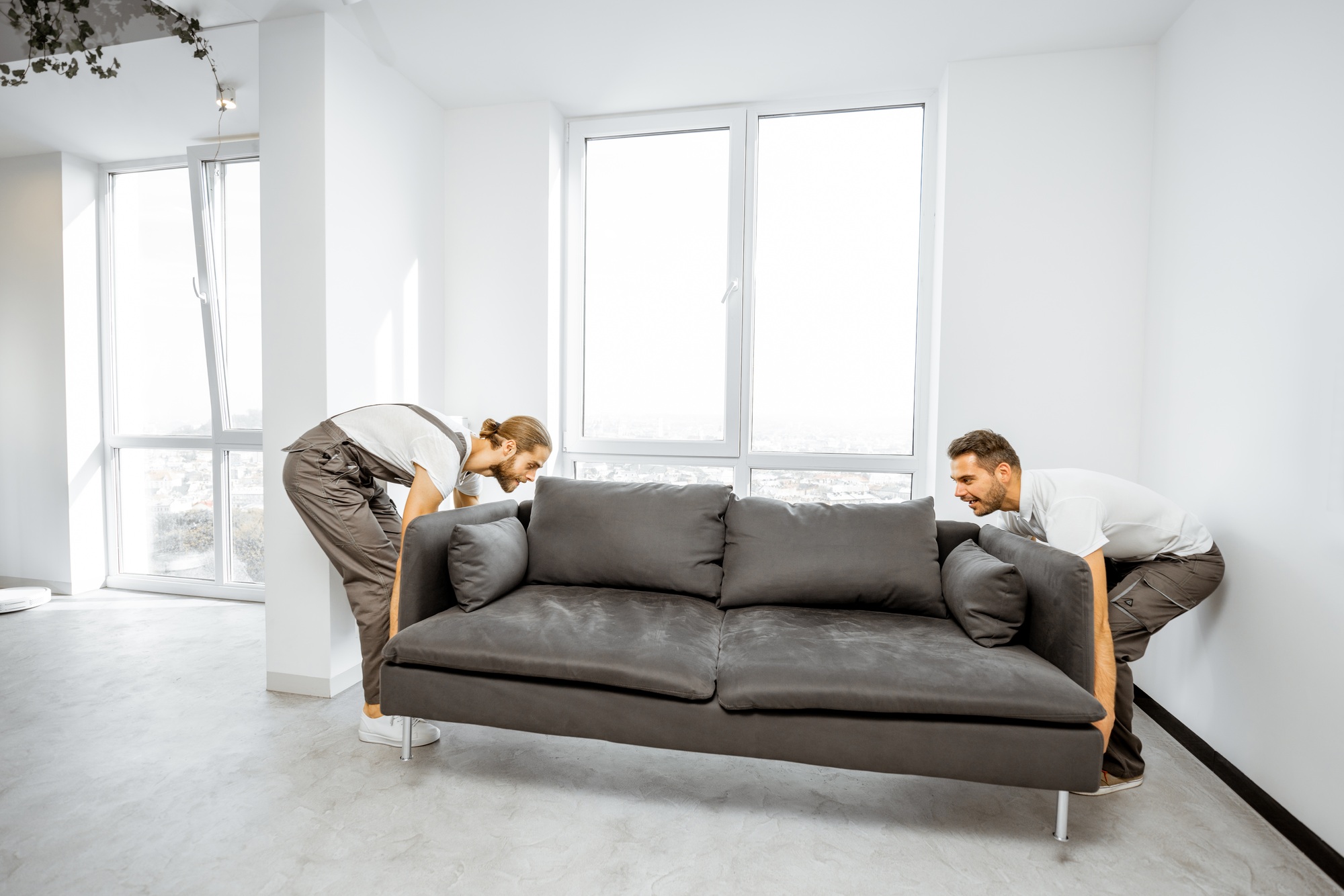 Deux hommes déplaçant un canapé gris dans un salon lumineux, près de grandes fenêtres. Déménager avec enfants conseils pratiques intégrés.