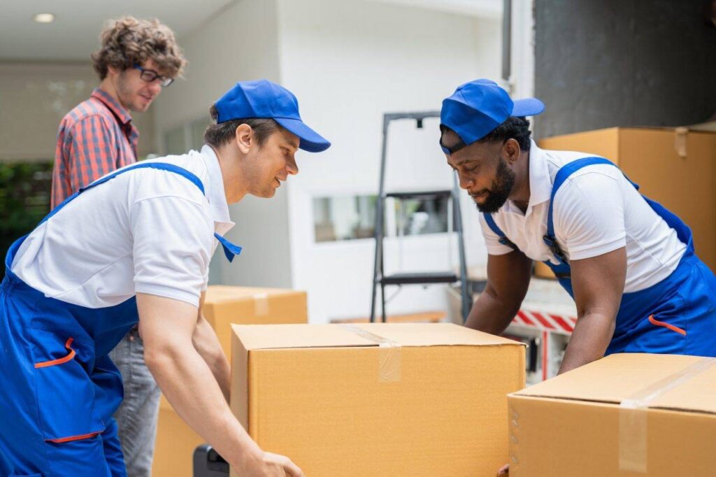 Deux déménageurs en uniformes bleus déplacent des cartons volumineux; un homme à l'arrière observe. Alt: aide de la caf pour déménager? (intègre le mot-clé).