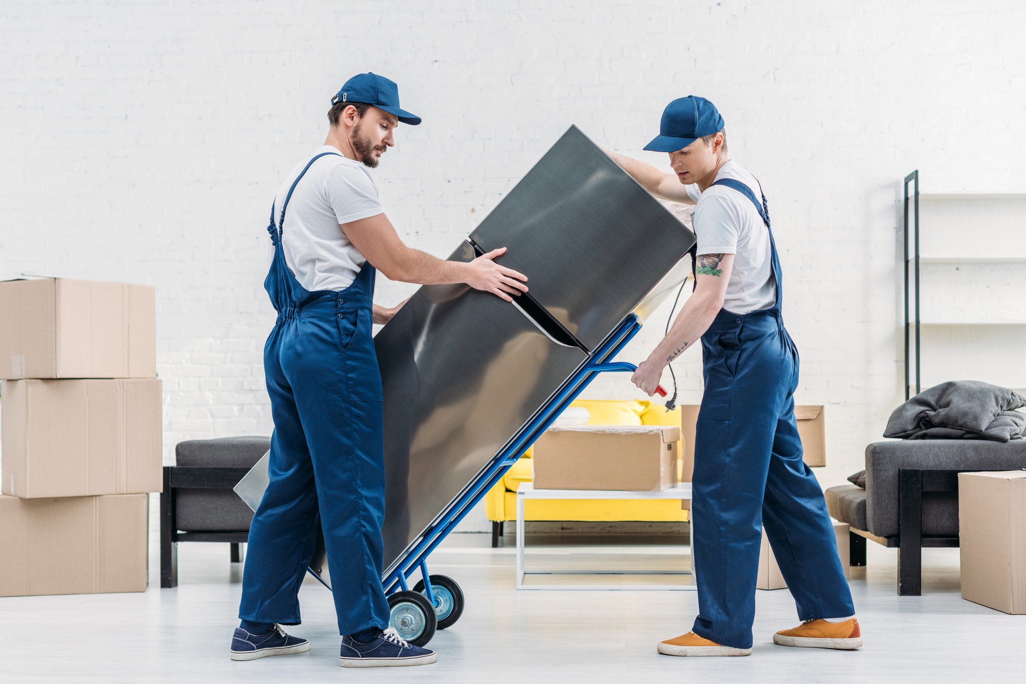Deux hommes en blue jeans et casquettes déplacent un grand meuble sur un diable, entourés de cartons et de mobilier. aide de la caf pour déménager.